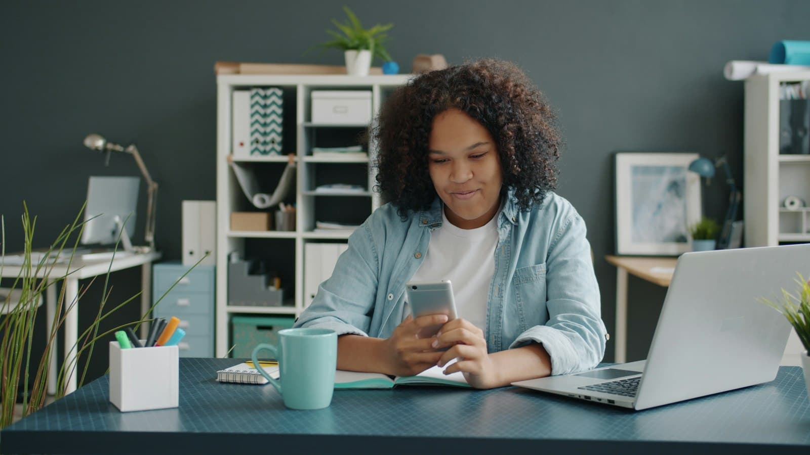 A freelancer at her desk, laptop open, checking a payment notification on her phone